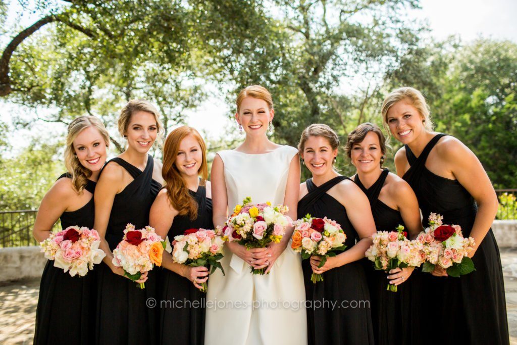 Bride in white with six bridesmaids in black gowns under live oaks, each holding a bright bouquet
