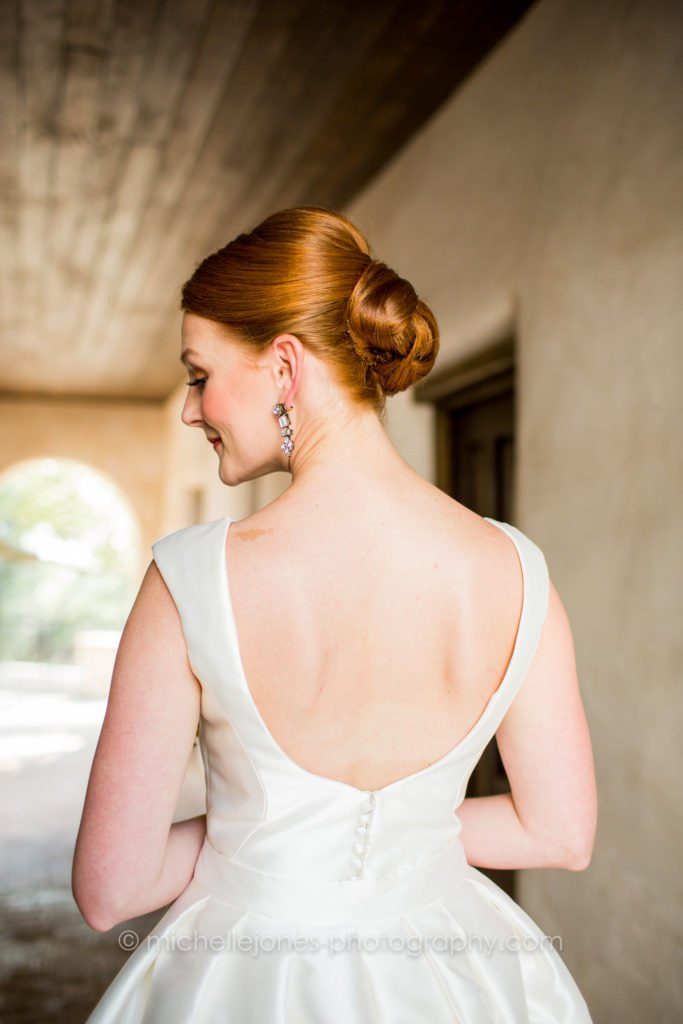 Red-haired bride photographed from behind, hair in a sleek French twist chignon, low-back satin gown