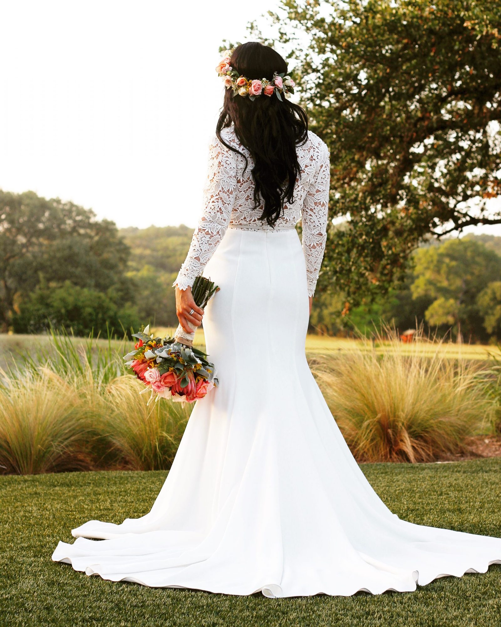A Chairish the Day bride from behind — loose dark waves under a fresh flower crown, lace sleeves, Hill Country light