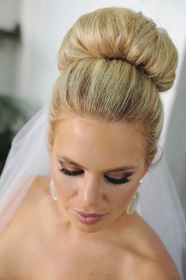 Bride with a polished high top-knot bun, lashes and a soft pink lip, veil behind